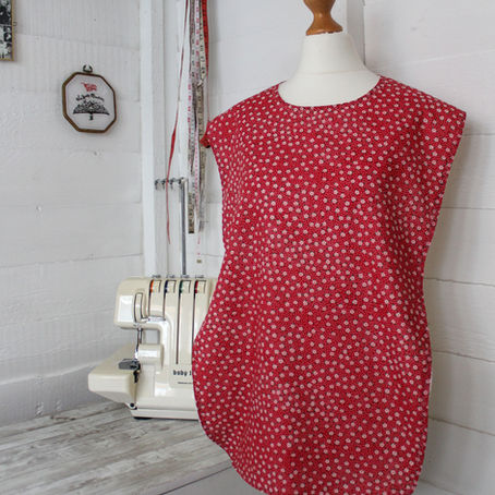 Mannequin in a sewing room wearing a red, patterned bib. Background has a sewing machine, tape measure, and framed decor. Cozy vibe.