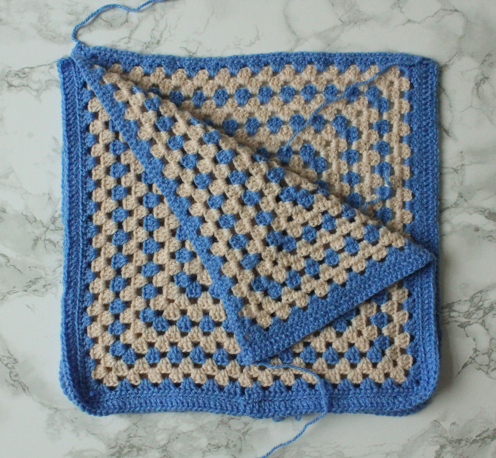 Crocheted blue and cream granny squares with folded corner on marble background. Visible yarn tail, intricate pattern, cozy mood.