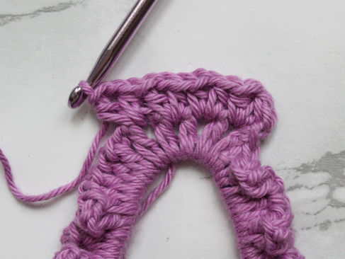 Purple crochet scrunchie in progress with metal hook on marble surface. The yarn is looped, showing a partially crocheted section.