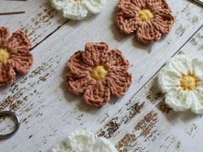 Crocheted flowers in white and pink with yellow centers on a rustic wooden table. Yarn, crochet hooks, and scissors are scattered around.