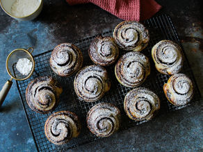Freshly baked chocolate and hazelnut brioche swirls dusted with powdered sugar on a cooling rack; sieve and bowl nearby with a red cloth in the background.