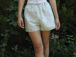 Young woman in a white tank top and floral shorts stands in a garden with greenery. She appears relaxed and is not displaying emotion.