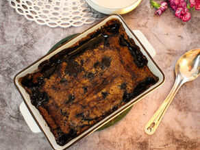 Baked pudding with a dark, crusty top in a white pan on a textured surface. Silver spoon and pink flowers nearby. Rustic ambiance.