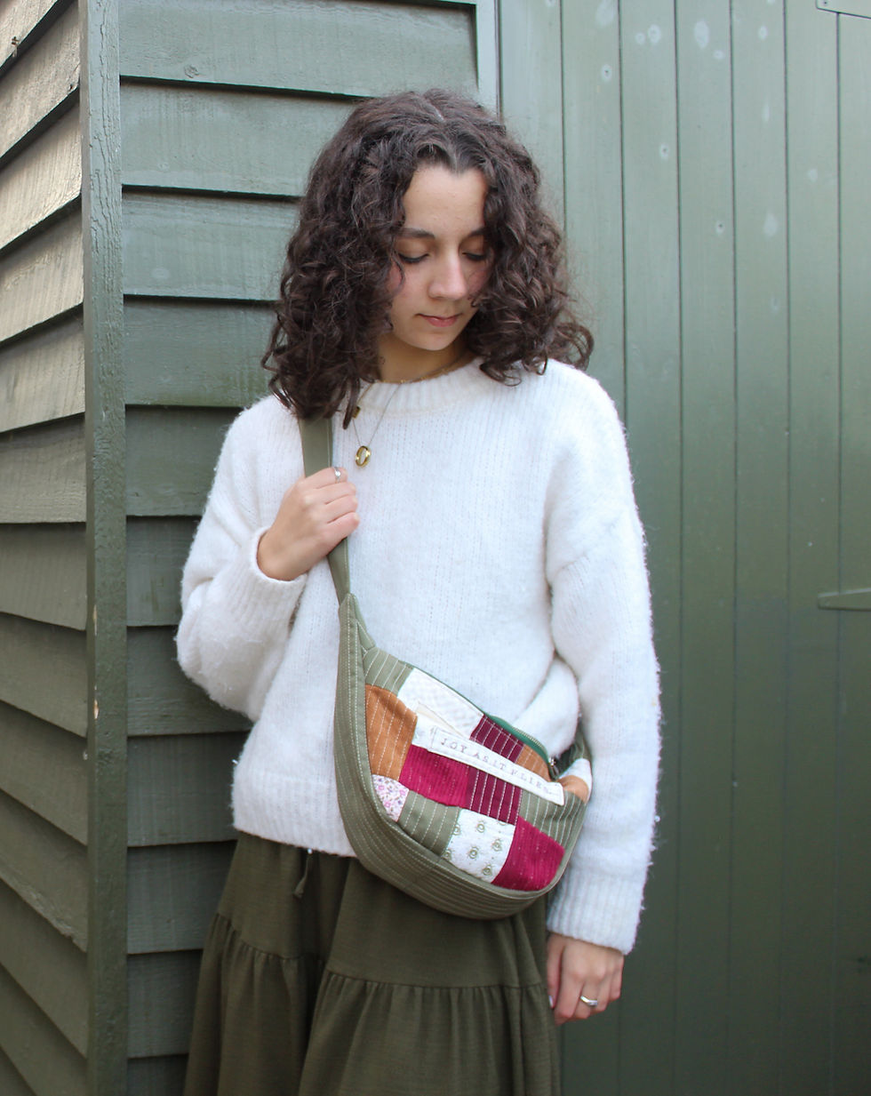 Young woman in a white sweater and green skirt, holding a colorful patchwork bag, leans against a dark green wooden wall, looking down calmly.