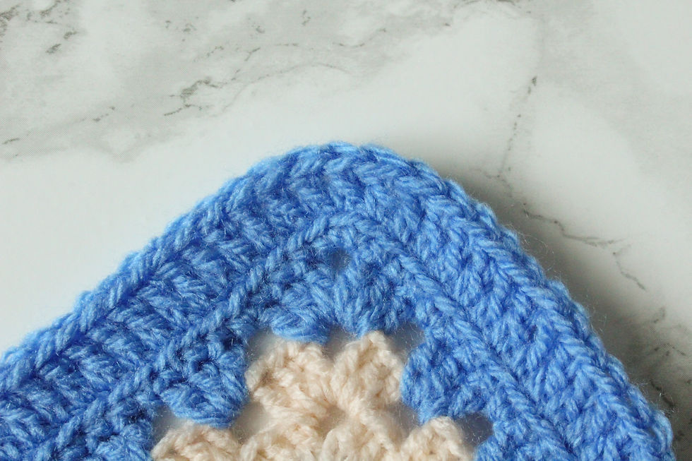 Close-up of a blue and cream crochet granny square on a marbled surface. The yarn is thick, with a detailed textured pattern.