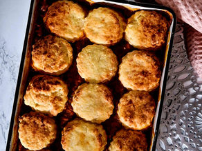 Baked dish with golden-brown biscuits on top, in a white pan. Red patterned cloth in the background. Warm, inviting kitchen setting.