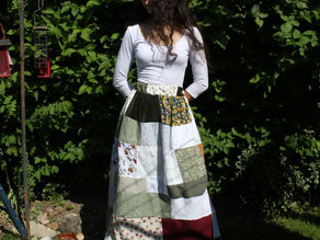 Woman in white top and patchwork skirt stands in a lush garden, beside red bird feeders; sunlight casts shadows, creating a serene mood.