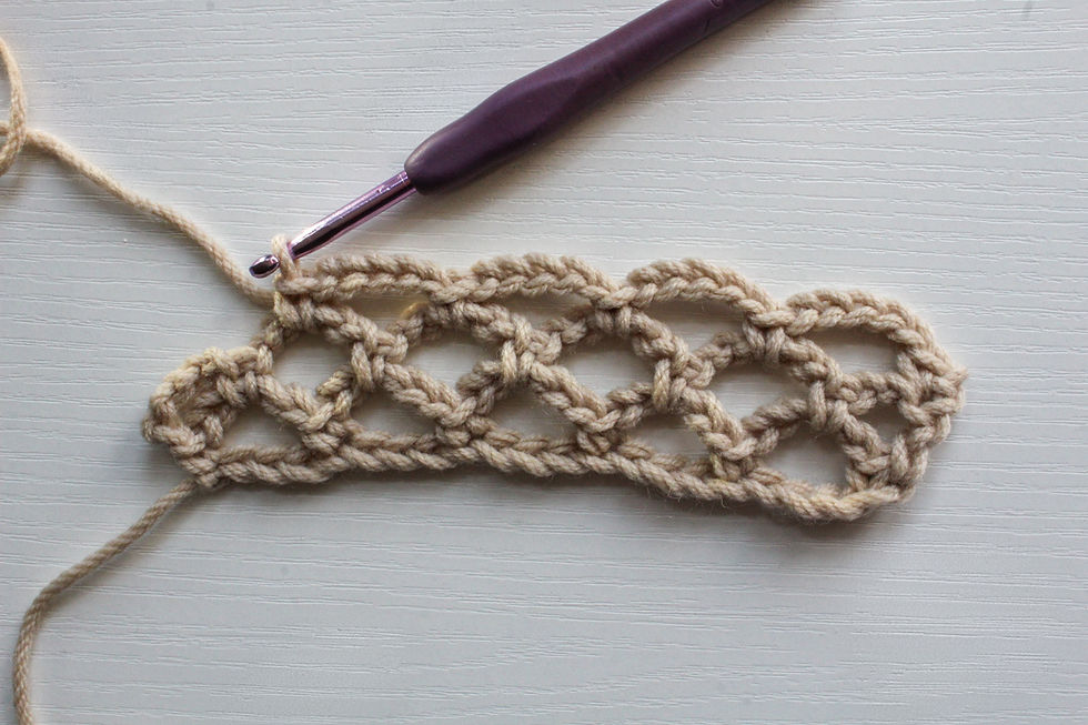 Crochet hook and beige yarn form a piece of crochet diamond mesh on a textured white surface. Hook is purple, yarn looped through stitches.