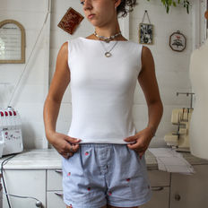 Person in a sewing studio holds the hem of a white top, wearing striped shorts, beside sewing machines and patterns.