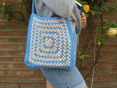 Young person with a crochet blue and beige bag holds a book, wearing a gray sweater and jean shorts. Brick wall and roses in background.