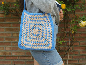Young person with a crochet blue and beige bag holds a book, wearing a gray sweater and jean shorts. Brick wall and roses in background.