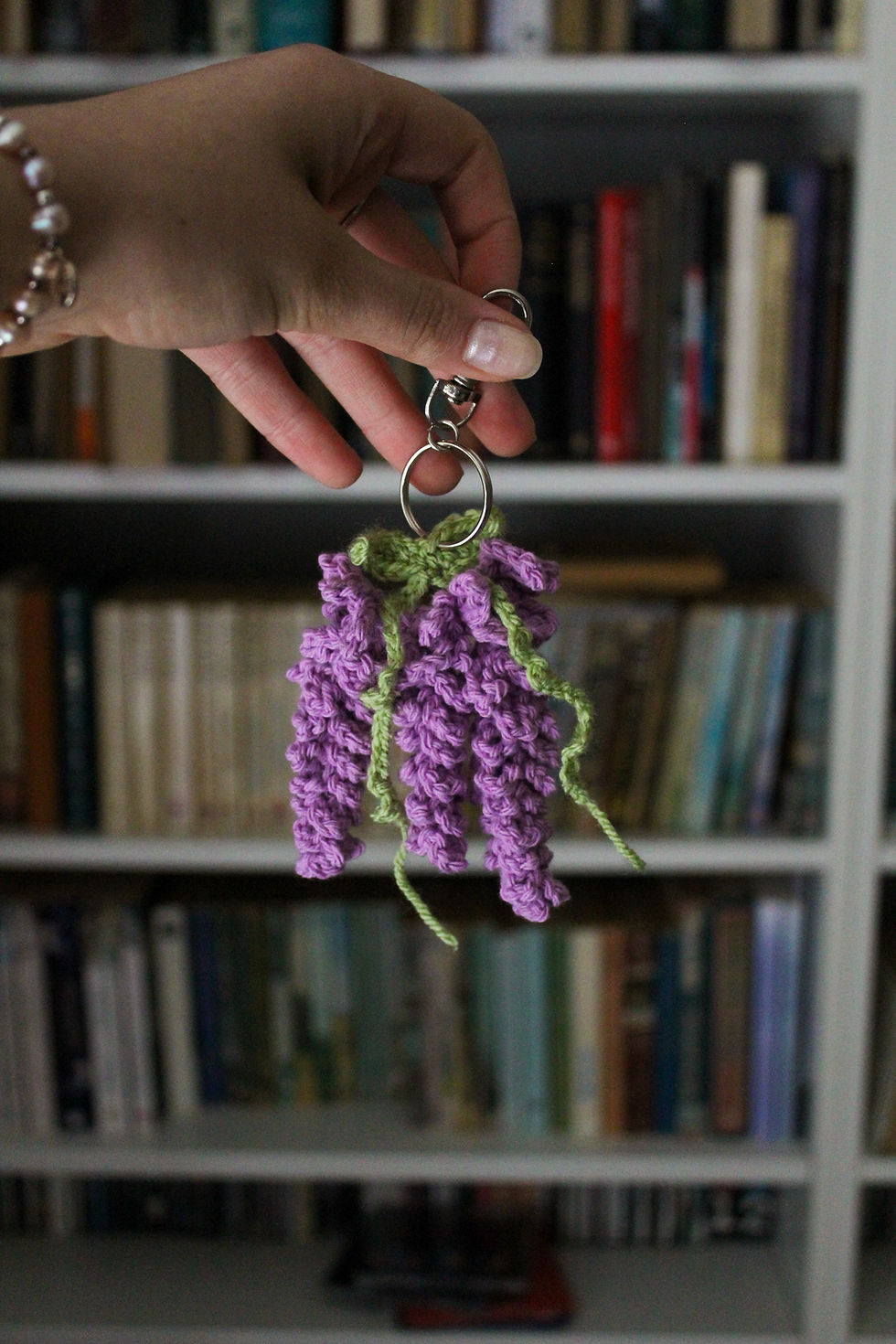 Hand holding a keychain with purple crochet wisteria flowers against a blurred bookshelf background, creating a cozy, crafty atmosphere.