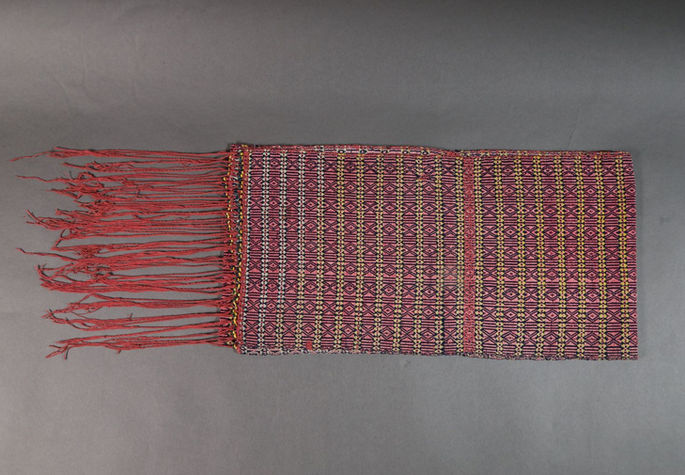 A hero shot of a rose-colored belt or loincloth folded in half with X and diamond motifs and braided fringes.