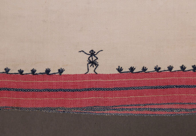 A detail shot of a human motif, alongside with what appears to be rice plants, embroidered on top of the red band border of the skirt