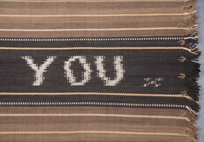A close-up shot featuring the word “YOU” woven into the textile, along with a shuttle-like motif, embroidery, and fringe
