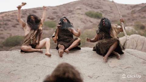 Three people dressed as cavemen cheer atop a rocky hill, displaying excitement. Dry, hilly landscape in the background.