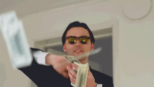 Man in suit and sunglasses joyfully throwing dollar bills indoors. White walls and door in the background. Energetic and lively mood.