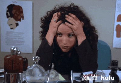 A person with curly hair holds their head in frustration, seated at a cluttered desk. Posters behind them. Logos for Seinfeld and Hulu appear.
