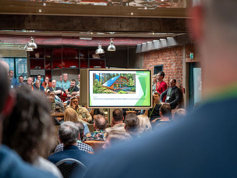 Menschen sitzen und stehen bei einer Veranstaltung und schauen auf einen großen Bildschirm mit Präsentationsfolie, Backsteinwand und Lampen im Hintergrund.