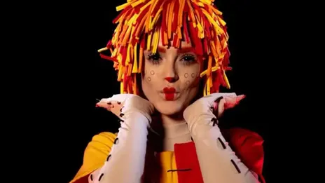 Isabelle Drummond caracterizada como Emília, com peruca laranja e amarela, figurino vermelho e amarelo e luvas brancas.