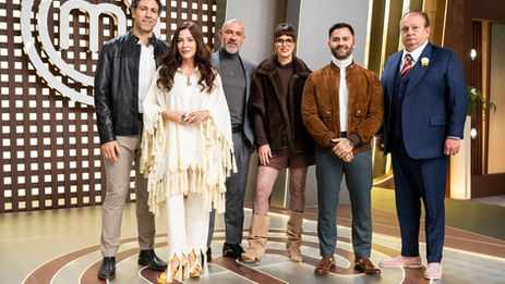 Seis integrantes e convidados do “MasterChef Profissionais” posam juntos no cenário do programa, em frente ao logotipo do MasterChef.