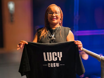 Lucy holds up a tee shirt which displays her name "Lucy" on the front. She smiles at the camera.