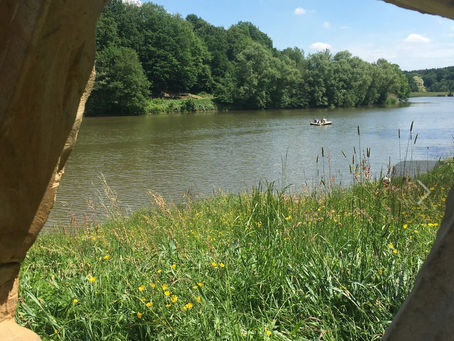 Blick durch eine Steinöffnung auf den ruhigen See mit einem Boot in der Ferne, umgeben von grüner Natur und gelben Blumen. Symbol für Reflexion, Loslassen und innere Ruhe.