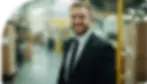 A smiling security guard in a black suit standing in a warehouse or factory setting. The background features blurred boxes and industrial equipment, indicating a secure working environment.