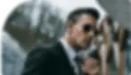 A security guard in a black suit, wearing dark sunglasses and an earpiece, holding a walkie-talkie, with a focused expression, likely monitoring a venue from a secure location.