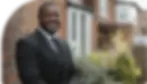 A security officer in a black suit, standing outside a residential area, possibly guarding vacant properties. He is smiling, with a well-groomed appearance.