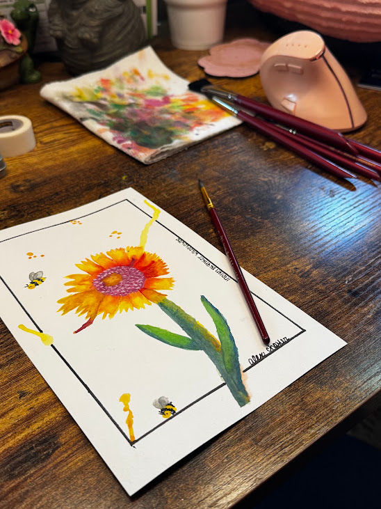 Watercolor sunflower painting on paper on a wooden desk, with brushes, a pink tool, and paint-smeared cloth nearby.