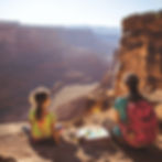 A sun-drenched photograph showing Alex Chavez as a child with her mother, sitting at the edge of a stunning canyon in New Mexico. They're perched on red rock overlooking vast desert landscapes with layered mesas stretching to the horizon. The young girl wears vibrant tie-dye clothes while her mother, wearing a turquoise shirt and carrying a pink backpack, sits beside her with an open watercolor set and paper between them. This moment captures the outdoor painting experiences that would later influence Alex's artistic style, with the dramatic Southwestern landscape serving as both subject and classroom for her developing artistic vision.