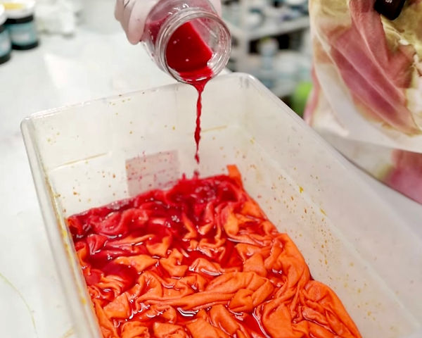 Corky is pouring orange dye from mason jar into a bin with a shirt being dyed.