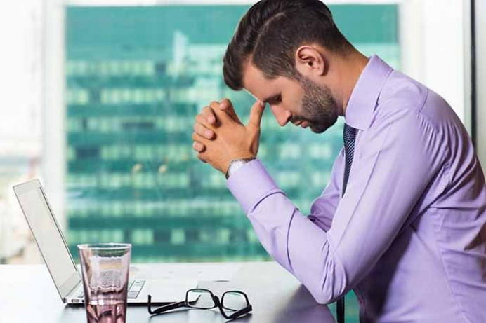 Man in purple shirt sits at desk with head down, appearing stressed. Laptop, glasses, and glass nearby. Office building visible outside.