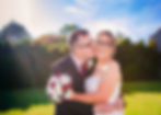 Bride and groom embrace outdoors; groom kisses bride's cheek. She holds a bouquet of red and white roses. Sunlight streams in; both smiling.