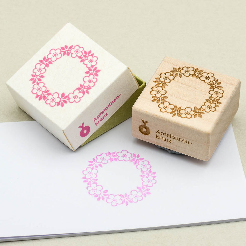 Holzstempel mit Apfelblütenkranz-Motiv, daneben eine bestempelte Karte und Verpackung. Design in Rosa und Grün.