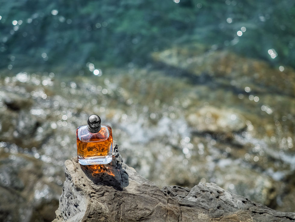 Fotografia commerciale di profumo su roccia con acqua sullo sfondo, luce naturale e riflessi per branding e e-commerce