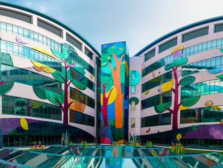 Colorful curved building on St Jude Children's Research Hospital campus with tree murals reflected in a glass courtyard pool under a blue sky.