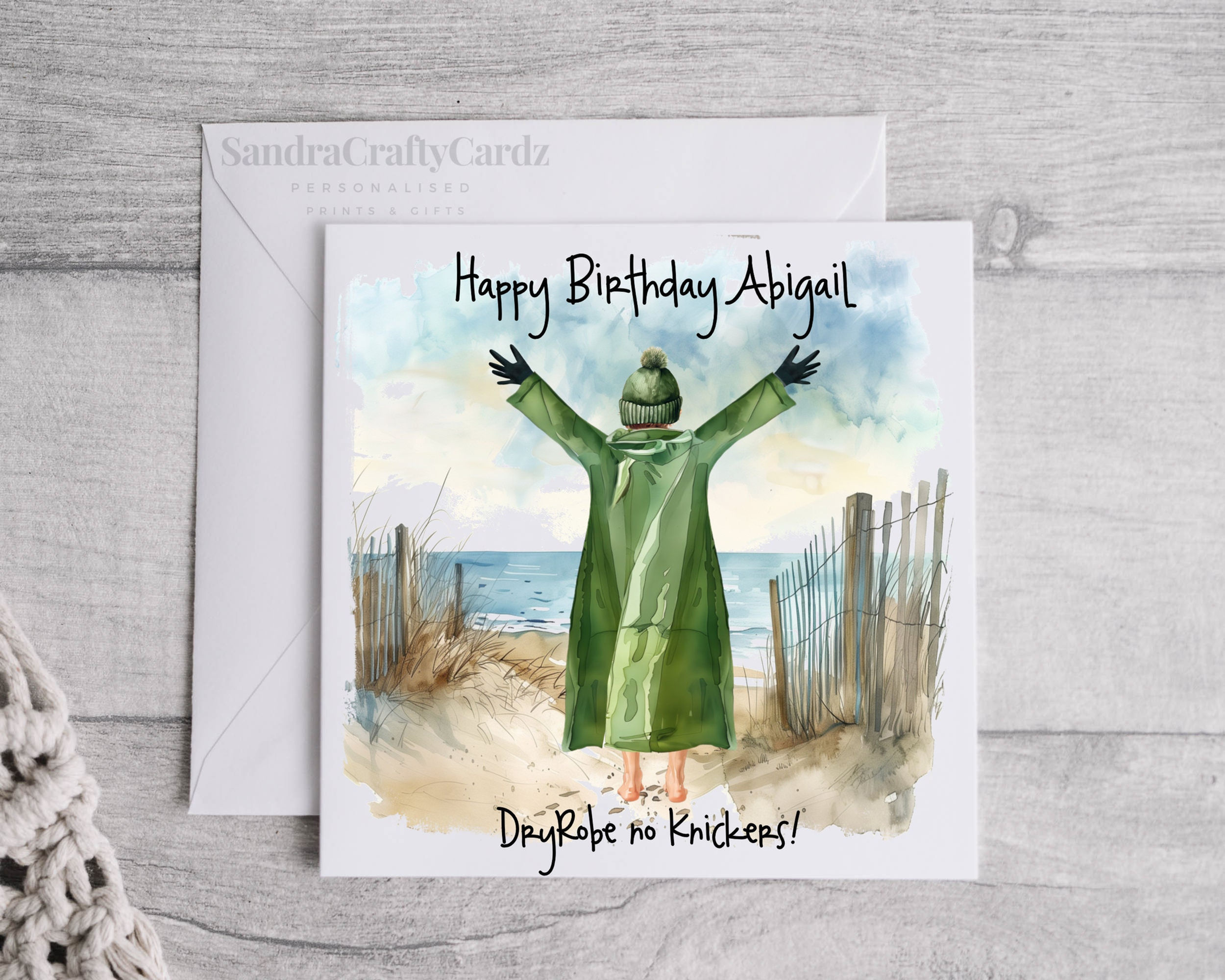 A personalised wild swimmer birthday card featuring a person in a green DryRobe and knitted hat standing on a sandy beach