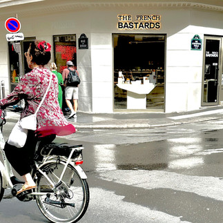 A woman cycling in a Parisian neighborhood with a baker in the background