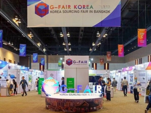 People exploring G-Fair Korea exhibit in a large, well-lit venue. Colorful banners hang overhead. Text: G-Fair Korea Sourcing Fair in Bangkok.