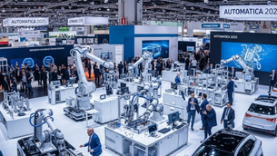 Robotic arms operate in a tech exhibition. Crowds in business attire engage around displays. "AUTOMATICA 2025" signs overhead. Futuristic mood.