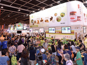 Crowded trade show with people browsing booths. Large white signs and screens display food items. Red "Exit Only" sign above. Busy atmosphere.
