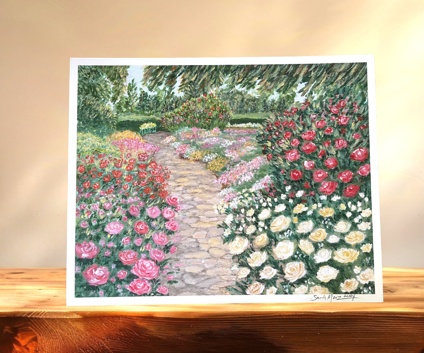 A painting of an english rose garden is displayed on a wooden table, backlit by natural light.