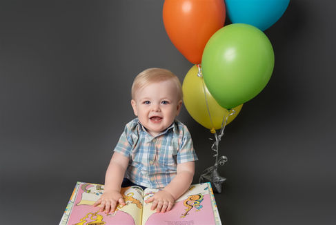 Little boy dressed in blue plaid holding a book and brightly colored balloons