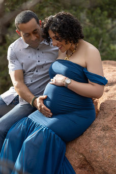 pregnant woman in blue gown sits on boulder with her partner as he touches her belly