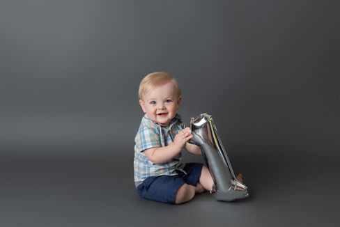 Little blonde boy dressed in blue holding a silver "1" balloon in front of a grey backdrop
