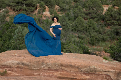 pregnant woman poses on boulder while her gown billows in wind