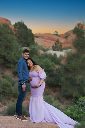 pregnant woman in lavender gown and husband in blue dress shirt and jeans pose for maternity photos against red rocks and trees