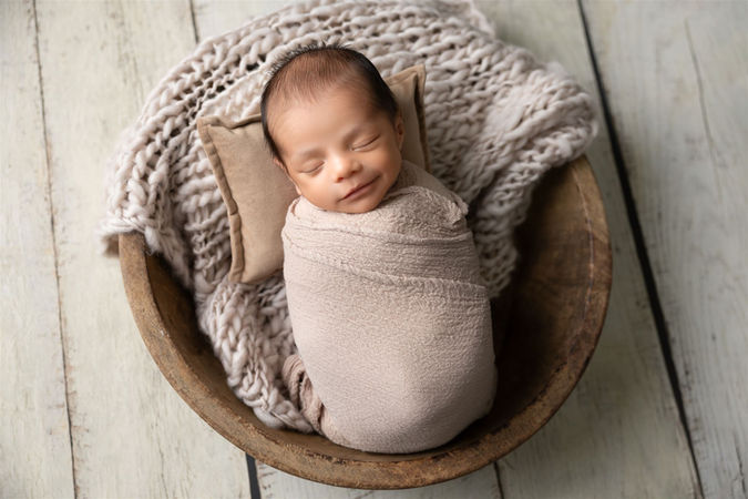 Newborn wrapped in tan fabric sleeps in bowl while smiling